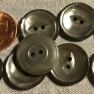 May include: Eight two-hole gray pearl buttons with a slightly iridescent finish. The buttons are arranged in a semi-circle on a gray fabric background.