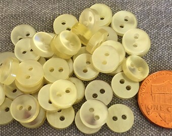 Könnte beinhalten: Eine Ansammlung hellgelber Knöpfe mit zwei Löchern. Eine US-Ein-Cent-Münze dient zum Größenvergleich. Die Knöpfe sind rund und haben einen leicht irisierenden Glanz. Die Knöpfe sind auf einer grauen Stoffoberfläche verstreut.