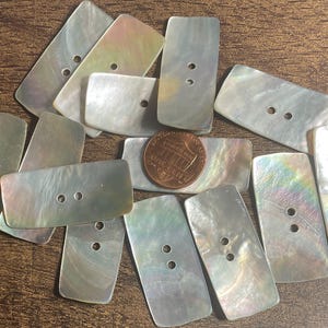 May include: A collection of rectangular mother-of-pearl buttons with two or three holes. The buttons have a pearlescent sheen, displaying iridescent colors. A U.S. one-cent coin is included for size comparison. The buttons are scattered on a brown surface.