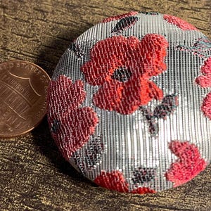 May include: A silver and red fabric button with a floral pattern. The button has a raised design and a shiny finish.