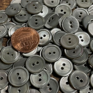 May include: A large pile of gray, two-hole, round buttons. The buttons are made of a pearlescent material and have a slightly iridescent sheen. A US penny is included for scale.