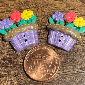 May include: Two decorative cupcake-shaped buttons with colorful flower accents. The cupcakes are purple with brown frosting and yellow polka dots. The flowers are red, yellow, and purple. A US one-cent coin is next to the buttons.
