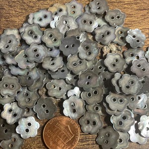 May include: A collection of iridescent, flower-shaped buttons with two holes each. The buttons are a mix of gray and silver, with a pearlescent sheen. A US penny is included for size reference.