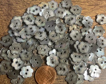 May include: A collection of iridescent, flower-shaped buttons with two holes each. The buttons are a mix of gray and silver, with a pearlescent sheen. A US penny is included for size reference.
