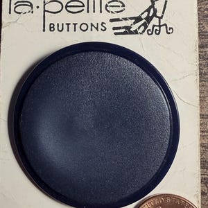 May include: A single dark navy blue circular button mounted on a cream-colored cardboard display card labeled la petite BUTTONS. The card includes the text 39 cents and 9389 70-1-3/4 inch. A United States one-cent coin is placed next to the button.
