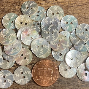 May include: A collection of iridescent, round buttons with two holes, each adorned with a delicate, white leaf design. A copper one-cent coin is included for size comparison. The buttons are scattered on a wooden surface.