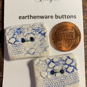 May include: Two white ceramic buttons with blue and white glaze. The buttons are rectangular and have a rough, textured surface. The buttons are machine washable. The text "The Logger's Daughter" and "earthenware buttons" are printed on the card. A US penny is included for scale.