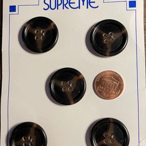 May include: A card with the word "SUPREME" printed on it. Four brown and black buttons with four holes each are displayed on the card. The text "4/$1.00 DEPT. 173" is printed on the card. A penny is also displayed on the card.