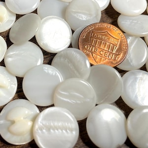 May include: A collection of white, round buttons with a pearlescent sheen, some with a decorative design. A copper one-cent coin is included for size comparison. The buttons are scattered on a brown surface.