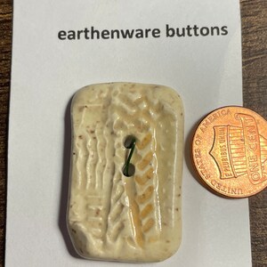 May include: A white ceramic button with a brown and tan design. The button is machine washable and is about the size of a US penny.