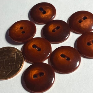 May include: Eight brown plastic buttons with two holes each. The buttons are slightly iridescent and have a glossy finish. The buttons are about the size of a US penny.