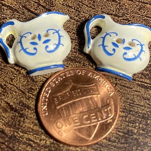 May include: Two small, cream-colored ceramic pitcher buttons with blue floral designs and blue trim. A copper penny with the words "One Cent" is next to the buttons. The buttons have a handle and a spout.