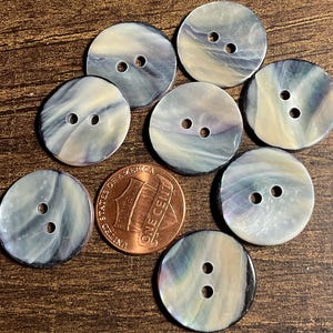 May include: Seven round, two-hole buttons with a swirling, iridescent gray and cream pattern. The buttons are arranged around a copper-colored United States one-cent coin, which serves as a size reference for the circular fasteners.