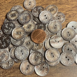 May include: A collection of round, gray buttons with a pearlescent sheen. Each button features two holes and a white, stylized floral design. A copper penny is included for size comparison. The buttons are scattered on a wooden surface.