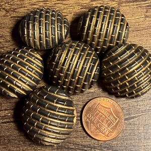 May include: Six round, dark brown buttons with a textured, grid-like pattern of raised ridges and recessed lines. A copper-colored United States one cent coin is positioned next to the buttons to provide a visual scale for their size.