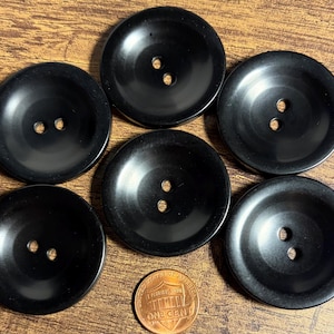 May include: Five circular black buttons with two central holes arranged in a cluster. Each button features a smooth, slightly concave surface. A copper-colored one-cent coin is positioned at the bottom center for scale, showing the Lincoln Memorial.