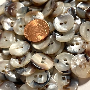 May include: A pile of white and brown shell buttons with a US penny for scale. The buttons are round and have two holes.
