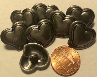 May include: A collection of antique silver-toned heart-shaped buttons. The buttons have a detailed, layered design and a loop on the back for sewing. A U.S. one-cent coin is included for size comparison. The buttons are likely for clothing or craft projects.
