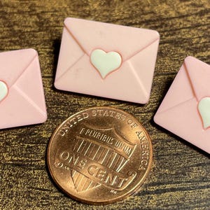May include: Three pink envelope-shaped embellishments with white heart accents, alongside a U.S. one-cent coin. The envelopes are arranged on a textured, brown surface. The coin is copper-colored.