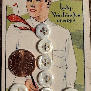 May include: A vintage card labeled Lady Washington Pearls featuring an illustration of a person in a white shirt and red tie, with seven white four-hole buttons attached. A United States one-cent coin is placed next to the buttons for scale.