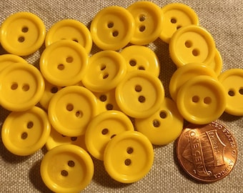May include: A selection of bright yellow, round buttons, each with two holes. A one penny coin is included for scale. The buttons are made of a smooth material and are scattered on a textured surface.