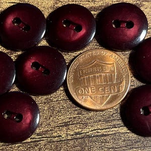 May include: A collection of nine dark burgundy buttons with two holes each, arranged around a copper one-cent coin. The buttons have a glossy finish and are round. The coin is from the United States.