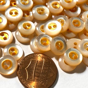 May include: A collection of 50 white pearl buttons with gold centers. The buttons are approximately the size of a US penny.