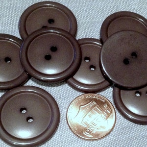 May include: A group of eight brown plastic buttons with two holes each. The buttons are arranged in a circle with a penny in the center.
