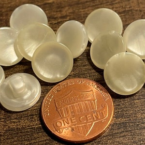 May include: A group of ten white, round, plastic buttons with a pearly sheen. The buttons are approximately the size of a US penny.