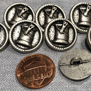 May include: Eight silver-toned buttons with a raised design of a spool of thread and a needle. The buttons are approximately the size of a US penny.