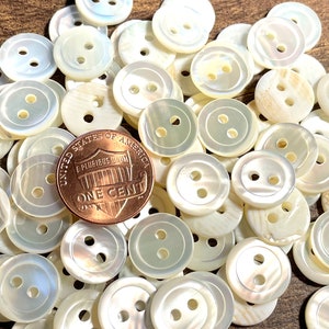 May include: A pile of white mother of pearl buttons with two holes each. A US penny is included for scale.