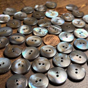 May include: A collection of round, two-hole buttons with a dark, iridescent, shell-like finish, scattered alongside two copper-colored one-cent coins. The buttons feature swirling gray and brown tones with a smooth, reflective surface texture.