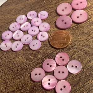May include: A collection of pink buttons with two holes each, scattered on a brown surface. The buttons vary in size and shade of pink. A copper penny is also present, providing a size reference for the buttons.