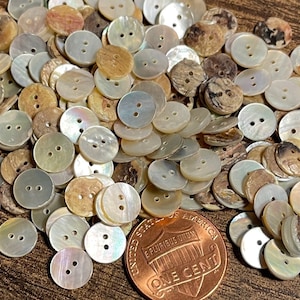 May include: A pile of round, two-hole, mother-of-pearl buttons in various shades of white, cream, and brown. The buttons are approximately the size of a US penny.
