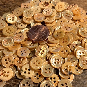 May include: A pile of small, round, beige buttons with four holes each. The buttons are made of mother of pearl and have a subtle iridescent sheen. A US penny is included for scale.