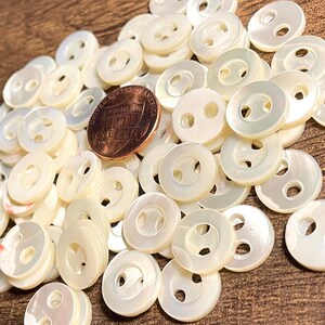 May include: A pile of white, iridescent, two-hole buttons. The buttons are made of mother of pearl and are approximately the size of a US penny.
