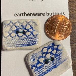 May include: Two white ceramic buttons with a blue and white pattern. The buttons are rectangular and have a hole in the center. The buttons are on a white card with the text "The Logger's Daughter" and "earthenware buttons" printed on it. The card also has the text "MACHINE WASHABLE" printed on it.