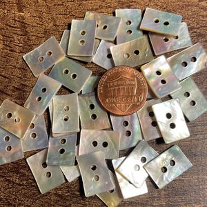May include: A collection of rectangular, iridescent mother-of-pearl buttons, each featuring two central holes. A copper one-cent coin is placed among the buttons to provide a sense of scale for these small, shimmering, off-white fasteners.