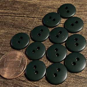 May include: A collection of 12 dark green plastic buttons with two holes each. The buttons are arranged in a semi-circle on a wooden surface.