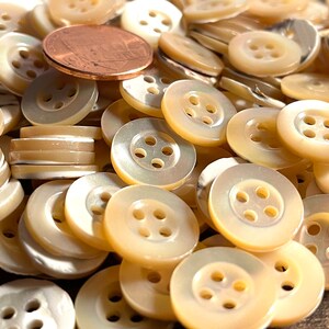 May include: A pile of approximately 100 white mother of pearl buttons with four holes each. The buttons are arranged in a random pattern and are all the same size. A penny is included for scale.