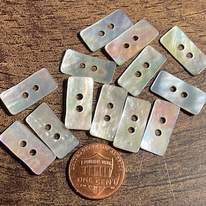 May include: A collection of rectangular, iridescent mother-of-pearl buttons, each featuring two centered holes. The buttons exhibit a shimmering, pearlescent finish with subtle color variations. A copper one-cent coin is included for scale.
