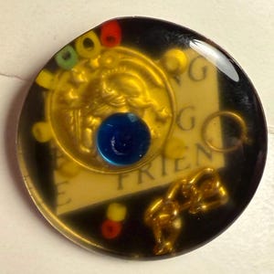 May include: A circular resin pendant featuring a gold-toned coin with a relief design, a central blue bead, and a gold-toned chain link embedded inside. The piece includes small red, yellow, and green beads, with partial text reading "G" and "FRIEN" visible on a light-colored background layer.