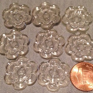 May include: Nine clear plastic buttons shaped like flowers with a raised center. The buttons are approximately the size of a US penny.