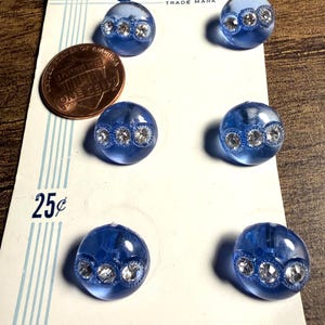 May include: Six round, translucent blue buttons arranged on a white Costumake brand card. Each button features a horizontal band of three embedded clear rhinestones. A copper-colored one-cent coin is placed next to the top-left button for scale.