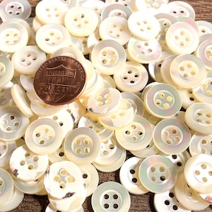 May include: A pile of white mother of pearl buttons with four holes each. The buttons are approximately the size of a US penny.