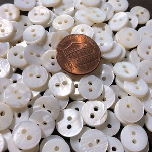 May include: A pile of white, two-hole, mother-of-pearl buttons. A US penny is included for scale.