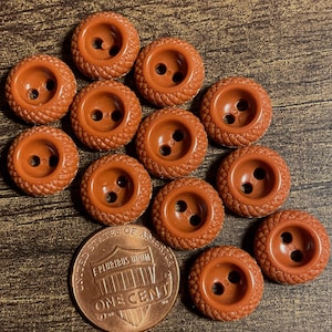 May include: A collection of eleven round, brown buttons with a textured, cross-hatched pattern on the outer rim and two center holes, displayed alongside a United States one-cent coin for scale to show the size of the small sewing fasteners.