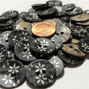 May include: A collection of black, oval-shaped buttons with a floral design and two holes. The floral design is white and the buttons are approximately 1 inch in diameter. A penny is included for size reference.