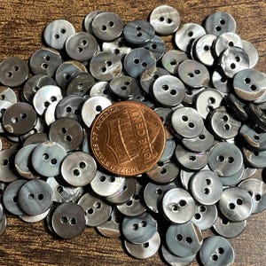 May include: A large collection of small, round, two-hole buttons with a grey, iridescent, marbled finish. A copper-colored one-cent coin is placed in the center of the pile to provide a sense of scale for the buttons.