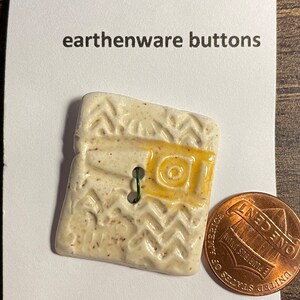 May include: A white ceramic button with a yellow and brown design. The button is machine washable and is shown next to a US penny for scale. The button is labeled "Logger's Daughter" and "earthenware buttons".